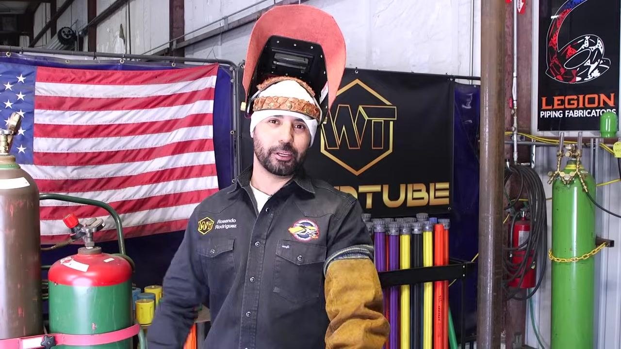 Instructor in welding shop, helmet up, WT jacket, American flag, WT banner, Legion Piping Fabricators poster, gas cylinders, colored pipe samples, and welding equipment visible. Delivering closing remarks.