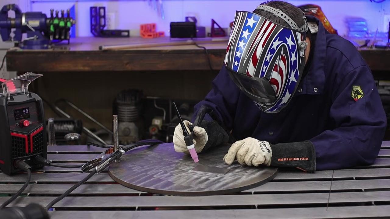 Welder in a protective jacket holding the TIG torch near the welder, with helmet and gloves ready on the bench.