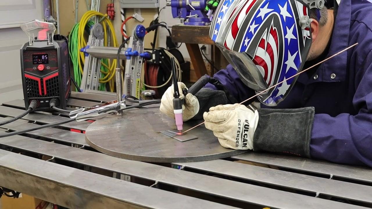 Welder in full safety gear performing TIG welding on a metal plate, with the welder display showing 85 amps and an organized workspace in the background.