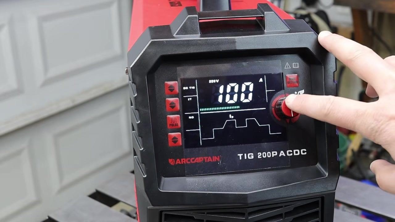 Close-up of the ARCCAPTAIN TIG 200P ACDC control panel showing 220V, 100A, waveform, and mode indicators, with a hand adjusting the red control knob and mode selection buttons visible.