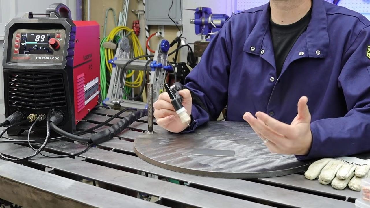 Person seated at a metal table with a red TIG welder showing "89" on the display, gloves, and welding accessories, with tools and helmets organized on a pegboard in the background.