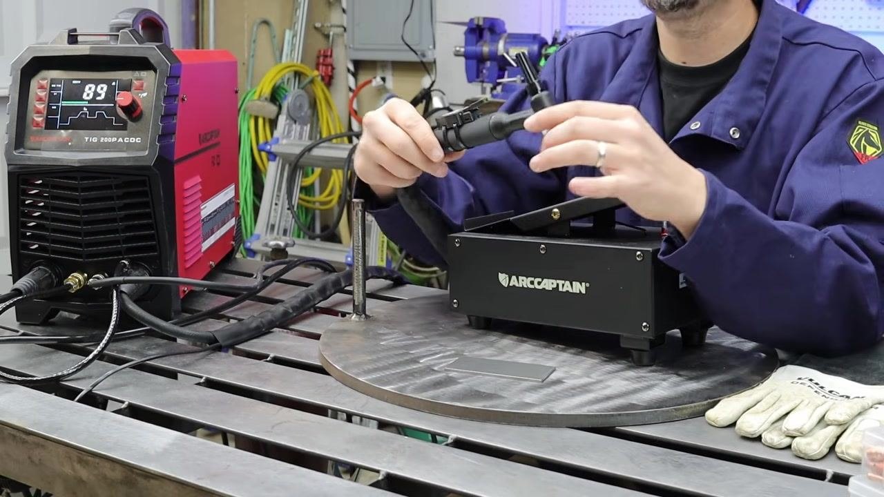 Person in a blue welding jacket demonstrating the TIG torch and ARCCAPTAIN foot pedal on a metal table, with the welder display reading "89" and gloves and tools organized in the background.