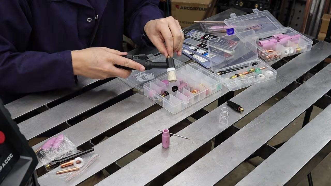 Person in a blue welding jacket preparing a TIG torch for a stubby gas lens upgrade, with organizer boxes of TIG accessories on the table.