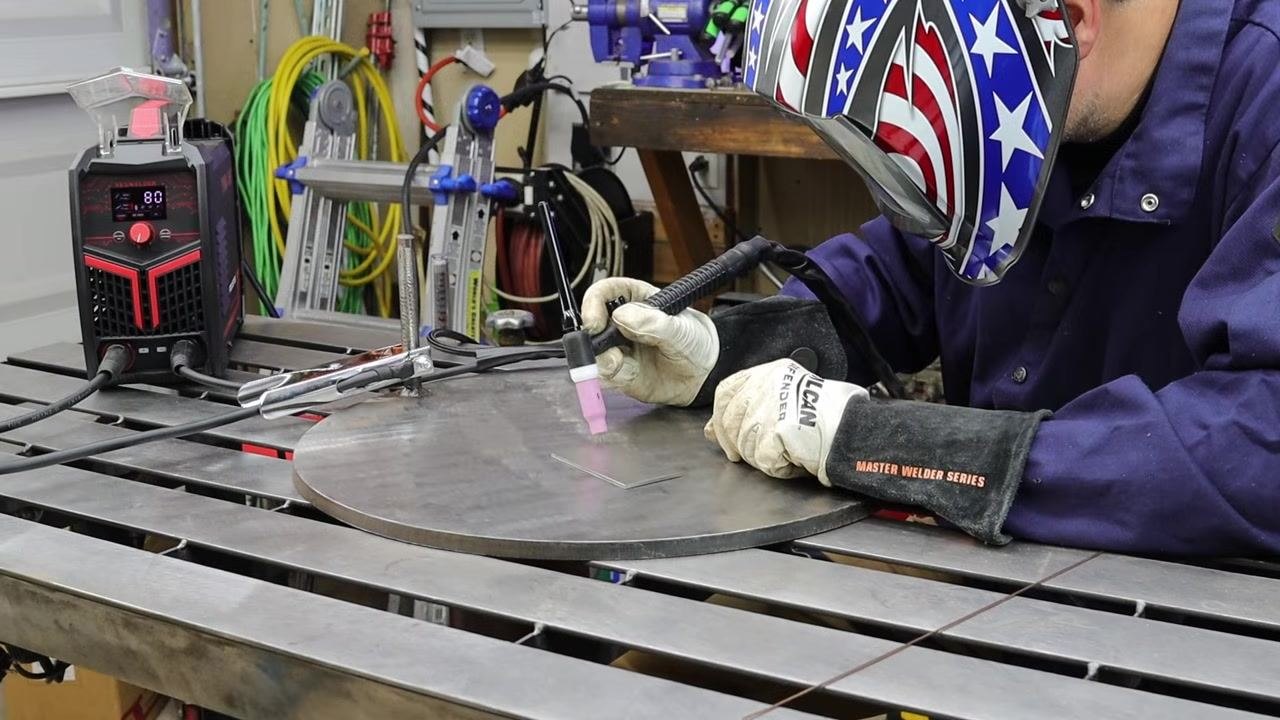 Welder using a stick machine with a TIG torch, wearing a welding helmet and gloves, preparing to weld a metal workpiece on a welding table with the machine set to 80 amps.