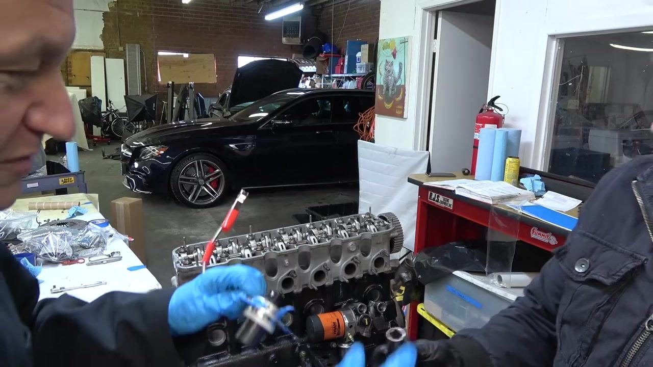 Person holding the angle gauge tool, preparing to attach it to the bolt, with a second person assisting. Engine and workspace visible.