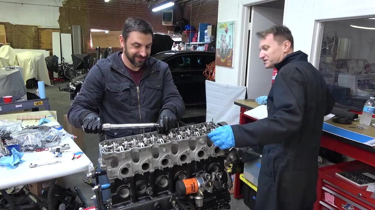 Person torquing the next cylinder head bolt following the cross pattern, with both individuals focused on the task. Engine and tools visible.