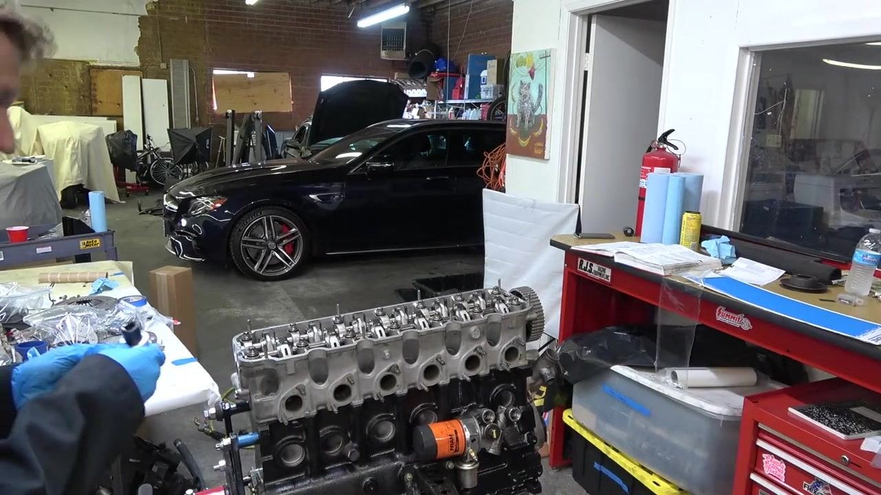 A person wearing blue gloves holds the angle gauge tool near the engine block, preparing for the 90° turn. The engine is mounted on a stand in a well-equipped garage, with a black car parked in the background and a red tool chest visible nearby.