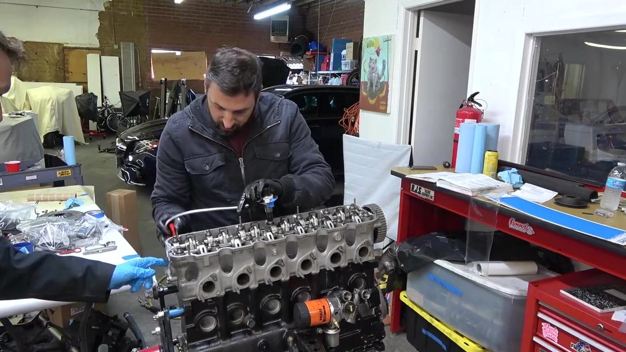 One person using a torque wrench with an angle gauge attached to a cylinder head bolt, while another assists. Workshop environment with tools and a car visible in the background.