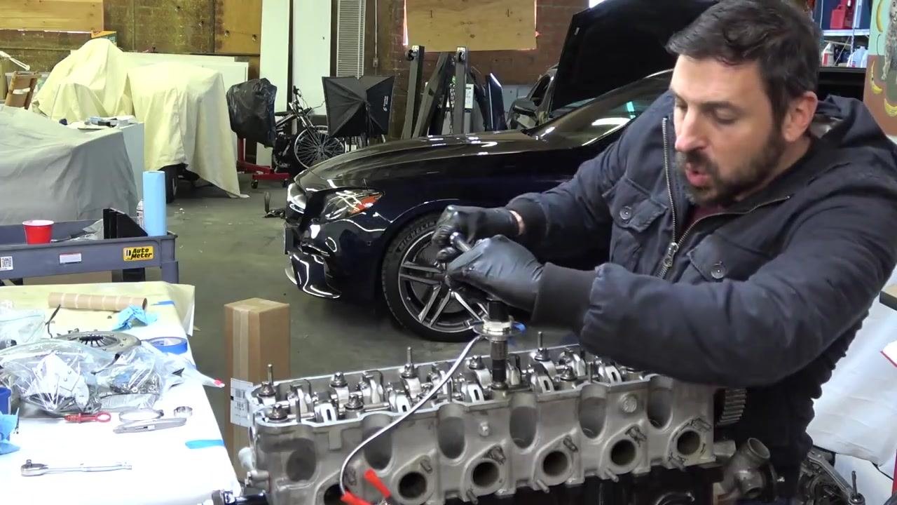 A person applies the final torque to the cylinder head bolt, focusing on the angle gauge and using controlled force. The engine, tools, and a car in the background are visible.
