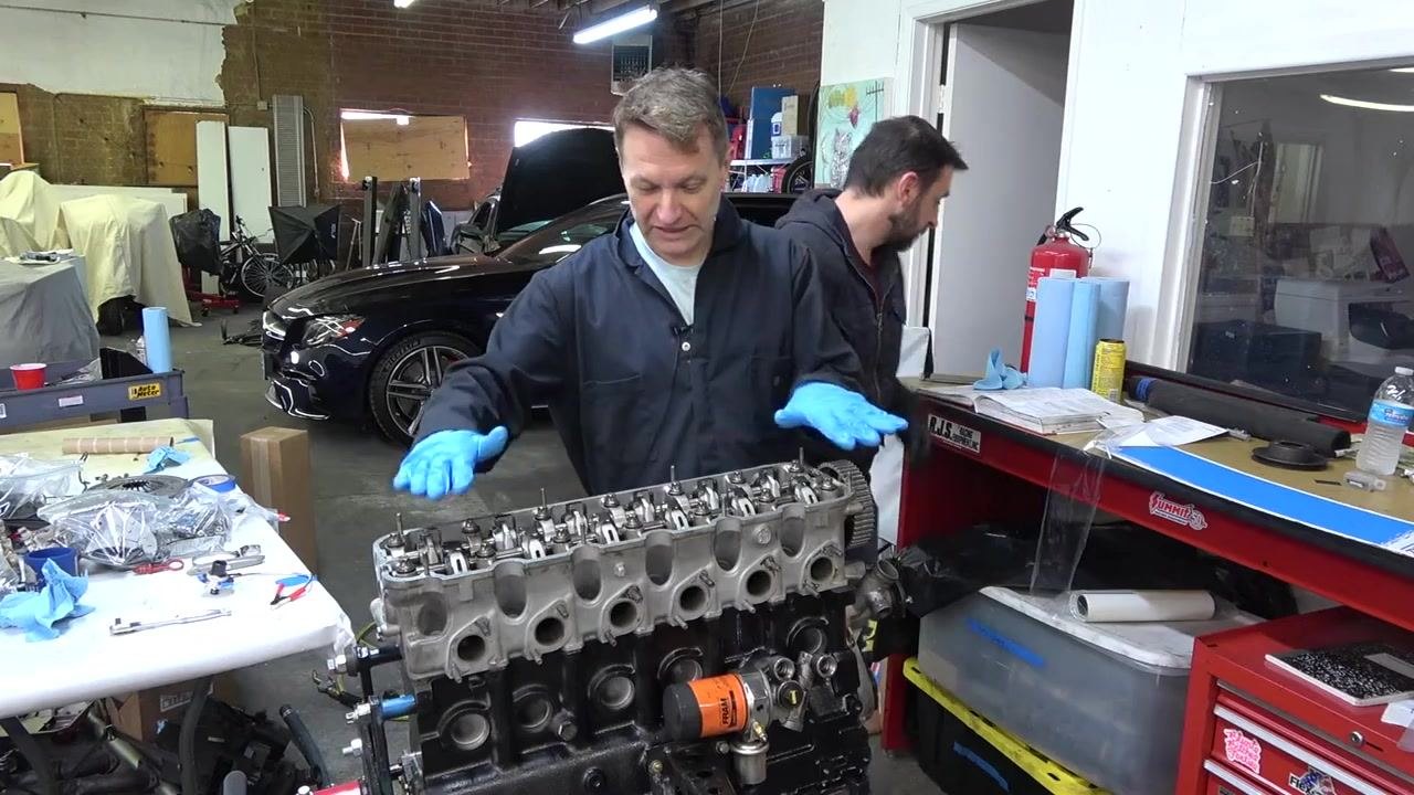 Person using a torque wrench to tighten the first cylinder head bolt on the engine, while a second person observes. Engine, tools, and workspace visible.