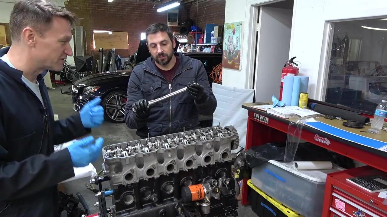 Two people in a garage: one holding a torque wrench and explaining its use while the other listens. The engine block, tools, and workbench are visible in the background.