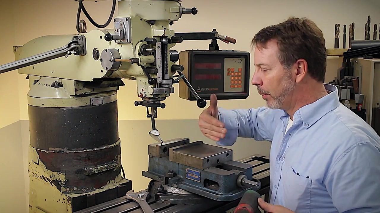 A machinist demonstrates vise alignment on a milling machine using a rotary base, a Mitsutoyo test indicator mounted on the spindle, a digital readout, and a soft-faced hammer, with the vise loosened and ready for adjustment.