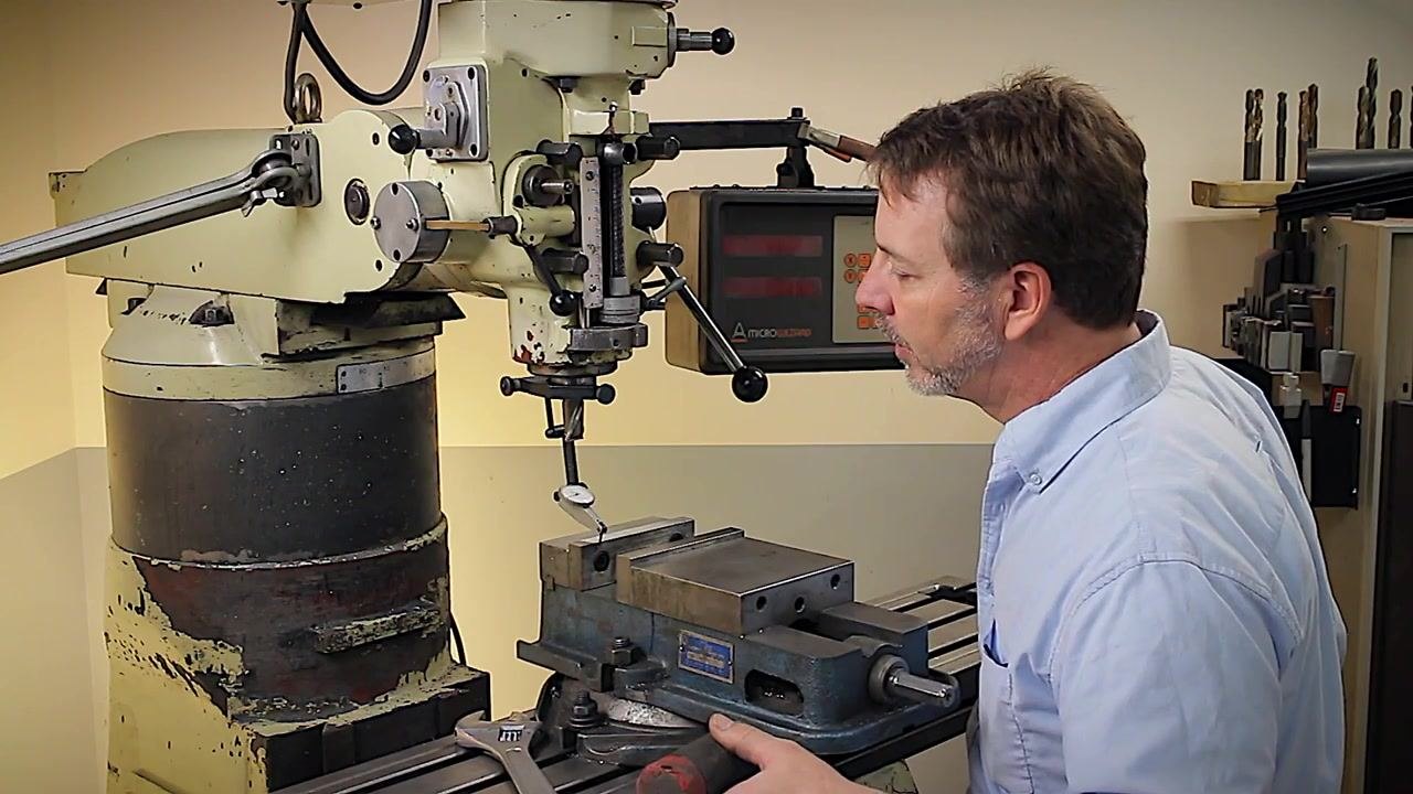 A machinist closely observes the test indicator while aligning the vise, with the indicator in contact with the fixed jaw and ready for fine adjustments, in a well-equipped machine shop with a digital readout and tools visible.