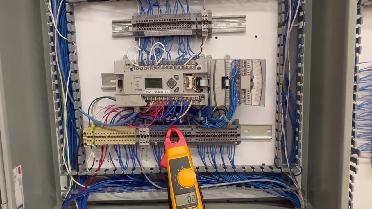 Control panel with the PLC and wiring visible, meter resting on the terminal block and displaying 0.0V, no probes held.
