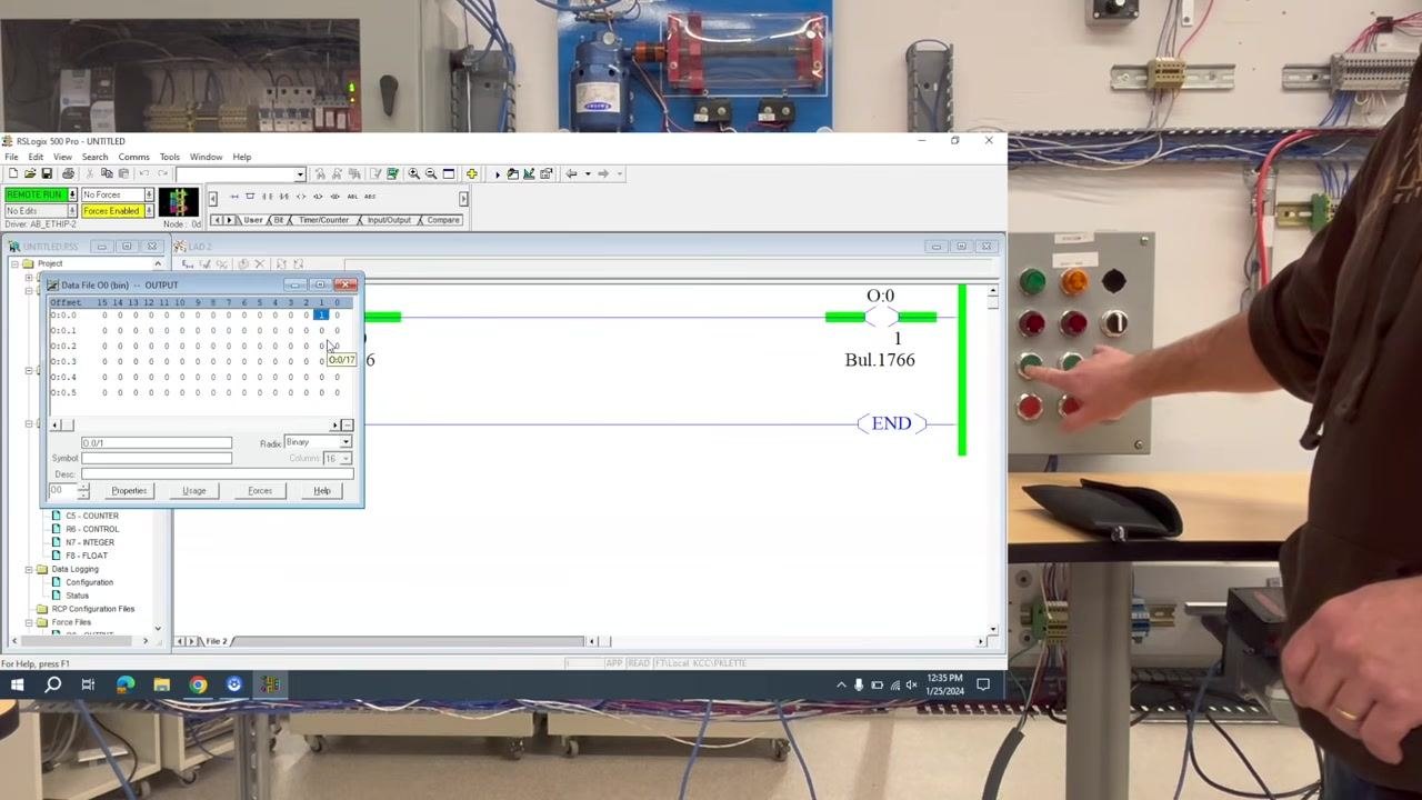 RSLogix 500 Pro software open on screen, showing ladder logic and the output data table while a hand presses the green push button on the control panel.