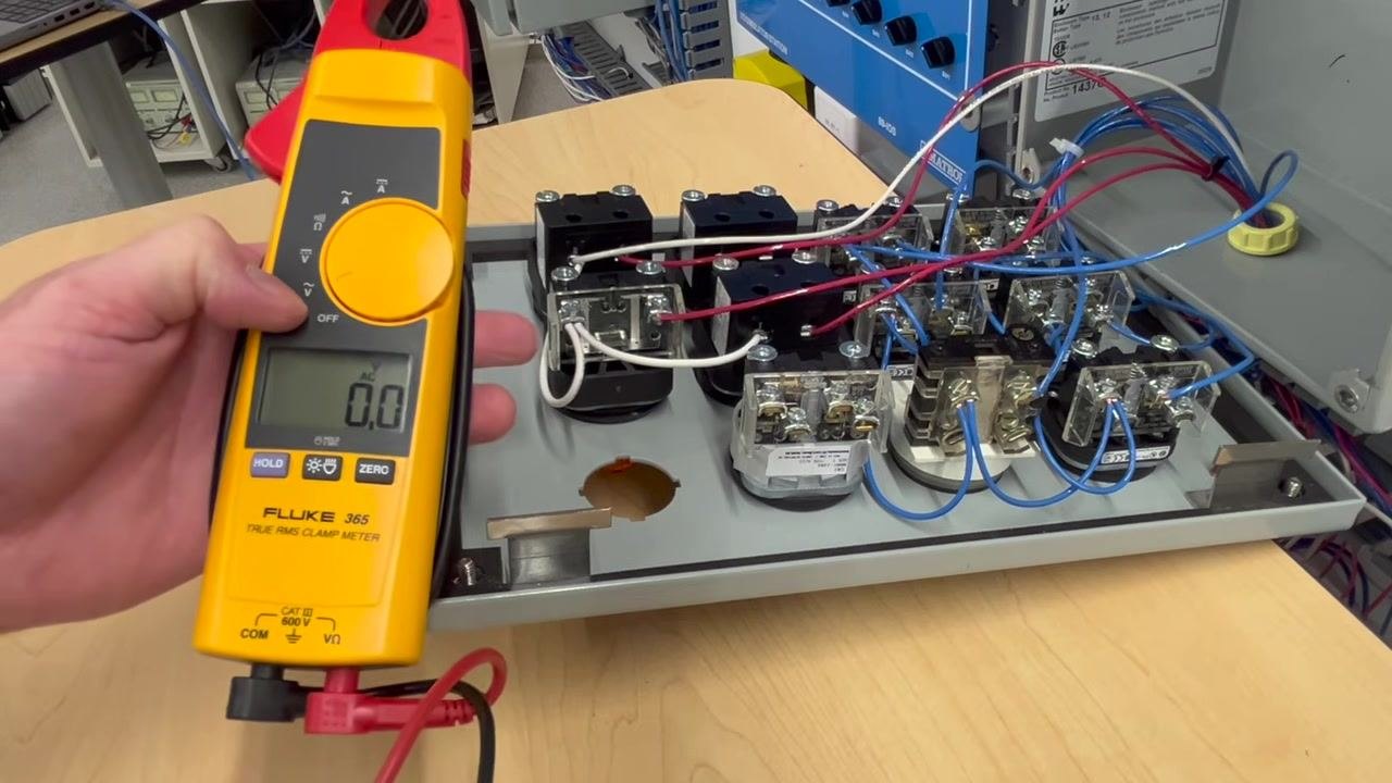 A hand holds a Fluke 365 True RMS Clamp Meter set to AC voltage, displaying 0.0V. The probes are near the output terminals inside an open control panel with red, blue, and white wires.