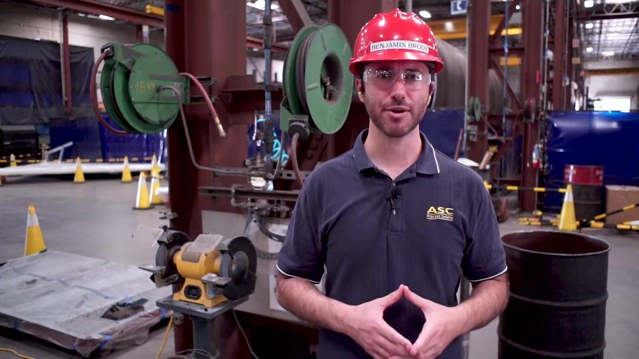 A worker in a hard hat and safety glasses stands beside a bench grinder in an industrial workshop, preparing to stop the wheel.