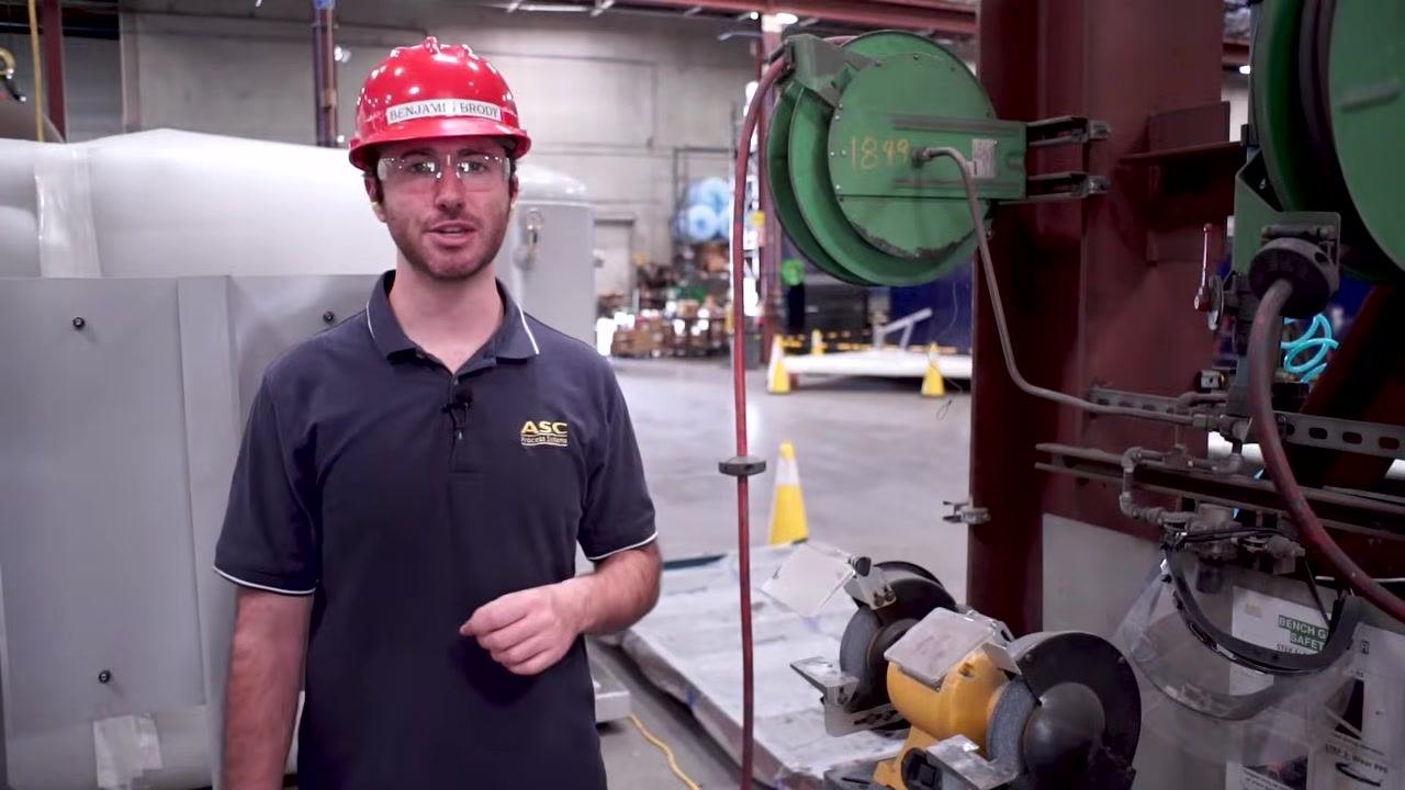 A bench grinder with safety tools attached to its pedestal, shown in an industrial environment with safety signage.