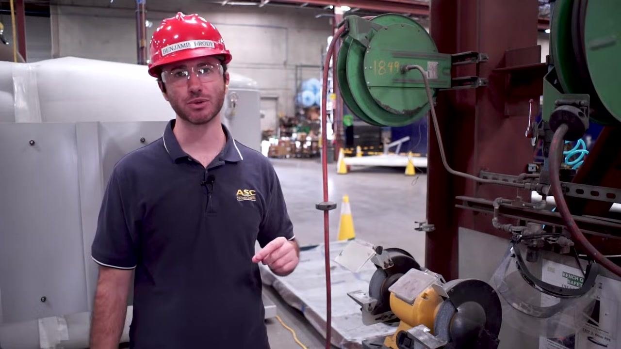 A worker demonstrates proper attire for grinding, wearing a fitted shirt, hard hat, and safety glasses in an industrial shop.