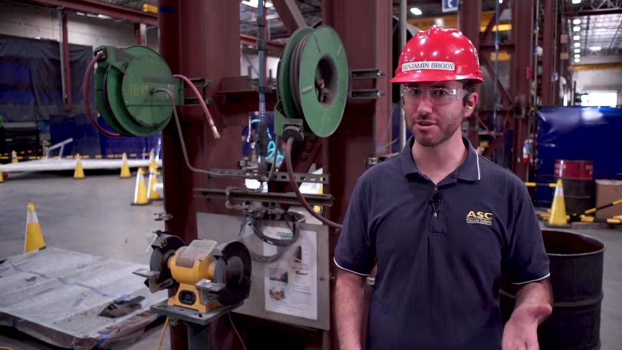 A worker equipped with a red hard hat, safety glasses, and ear protection stands beside a bench grinder, illustrating the required PPE.