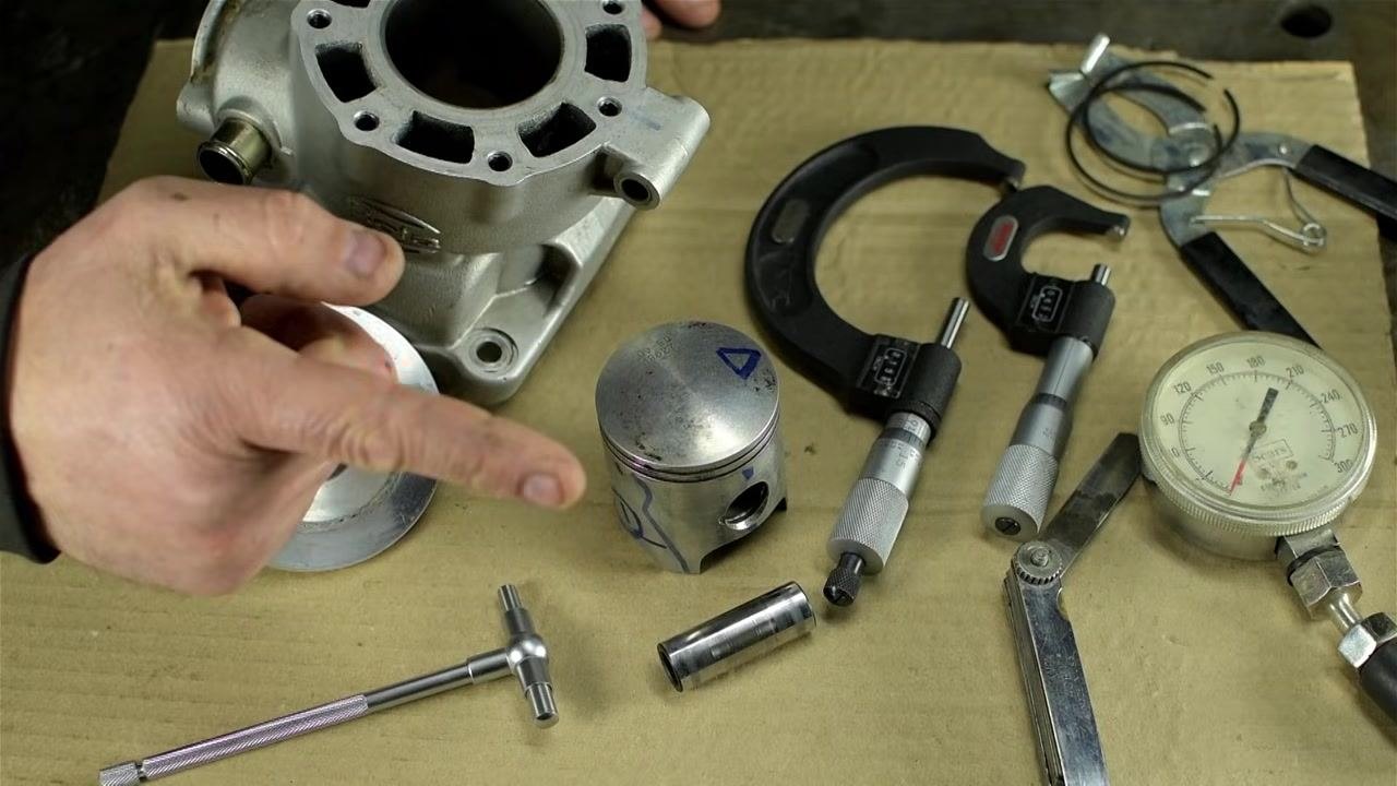 A workbench with a cylinder block, piston, piston pin, telescoping bore gauge, micrometer, dial indicator, feeler gauge, and calipers arranged on a cardboard surface, with a hand pointing at the piston.