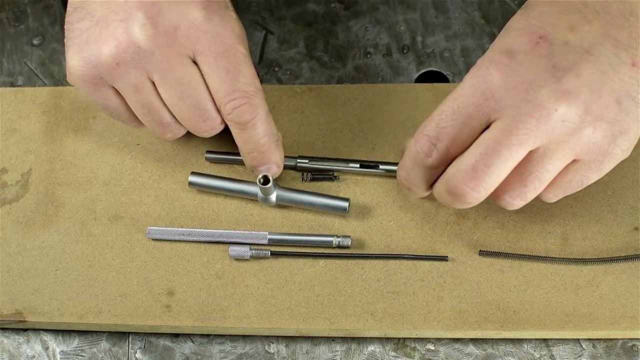 A close-up of several metal cylindrical parts, a spring, a threaded rod, and a hand holding one of the tubes, all laid out on a wooden work surface.