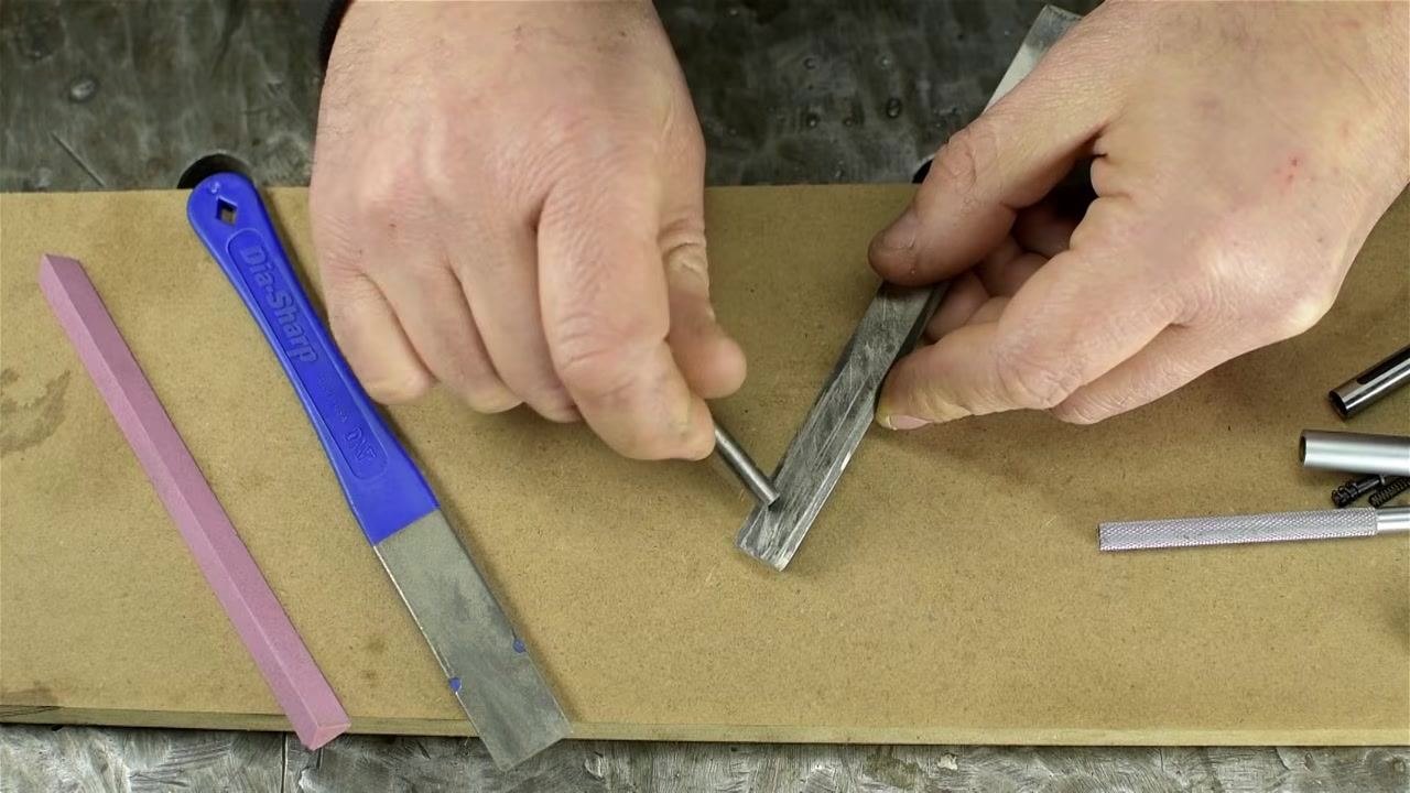 A hand uses a small cylindrical abrasive stone to deburr the inside of a metal tube while the other hand holds the tube steady, with other abrasive tools nearby.