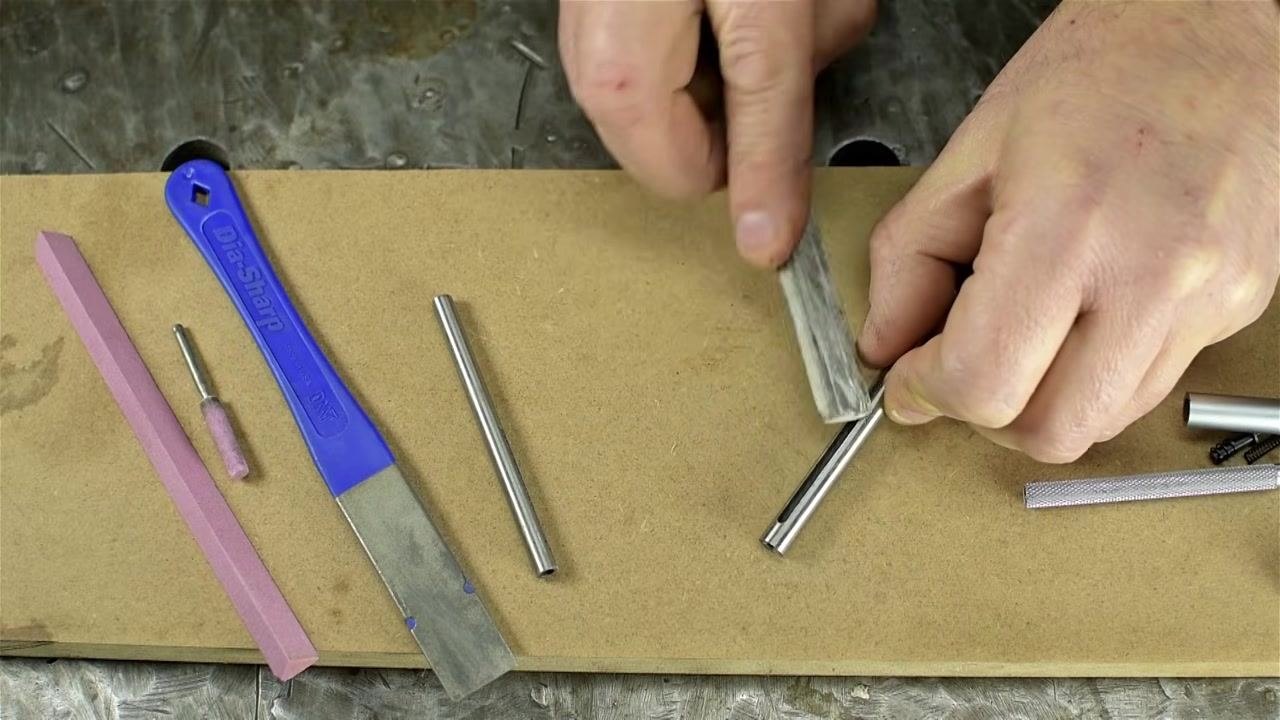 A hand uses a rectangular abrasive stone to deburr the edge of a metal tube while the other hand holds the tube, with other tools and parts visible on the work surface.
