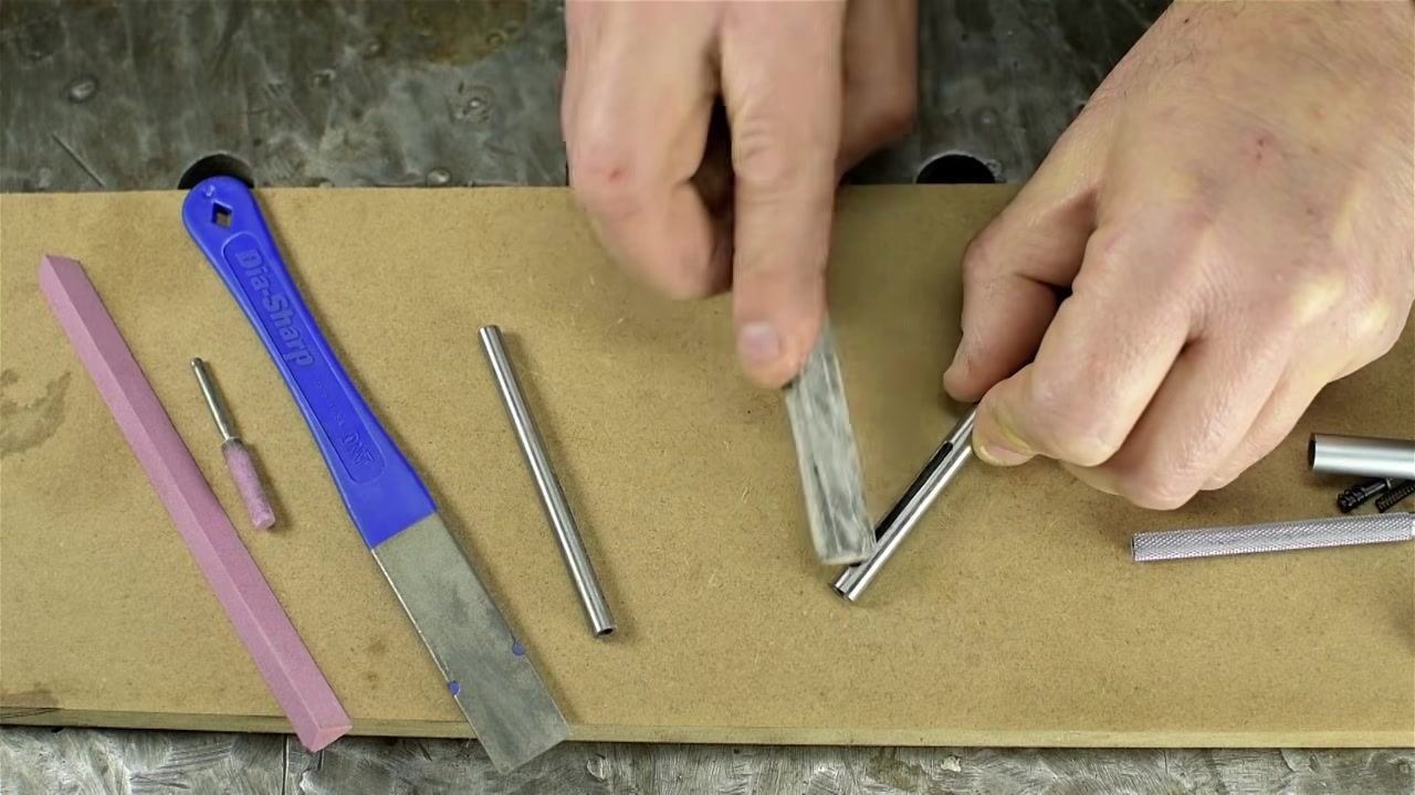 A hand continues to use the abrasive stone on the edge of a metal tube, ensuring all sharpness is removed.