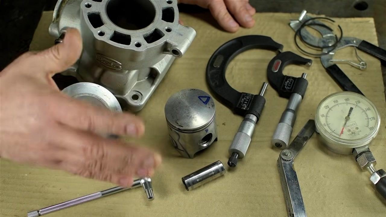 Close-up of a hand holding a telescoping bore gauge above a micrometer and piston, showing the gauge's construction and details.