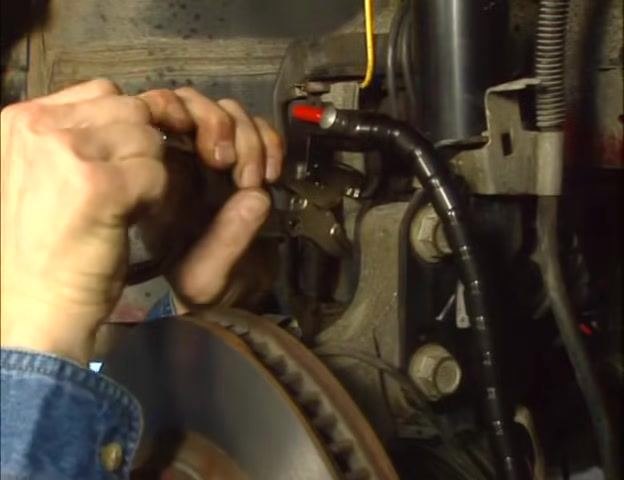Hands adjusting the knurled knob on a vise grip clamped to a vehicle suspension component, with a brake rotor visible in the foreground.