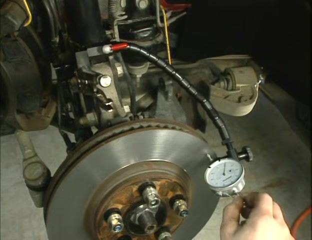 Person rotating the knurled ring at the end of the flex arm to remove slack, with the dial indicator positioned near the rotor.
