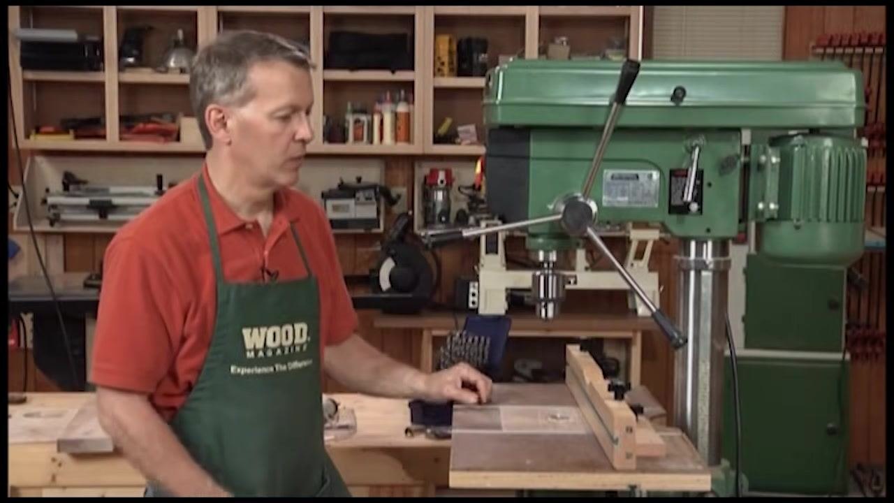 A person in a red shirt and green apron standing beside a drill press, discussing the benefits of using a drill press compared to a hand-held drill.
