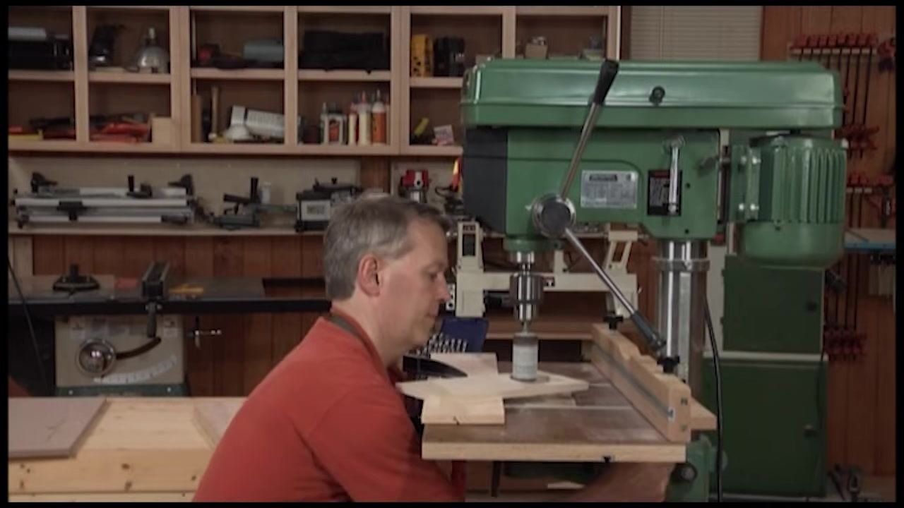 Hands positioning a wooden workpiece on the drill press table, with the sanding drum and wood dust visible