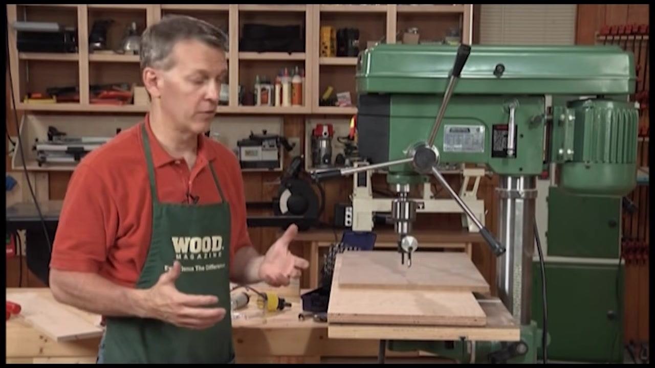 A person in a red shirt and green apron stands next to a green drill press, explaining its features. The drill press table has a large wooden board on it. The background shows a woodworking shop with shelves and tools.