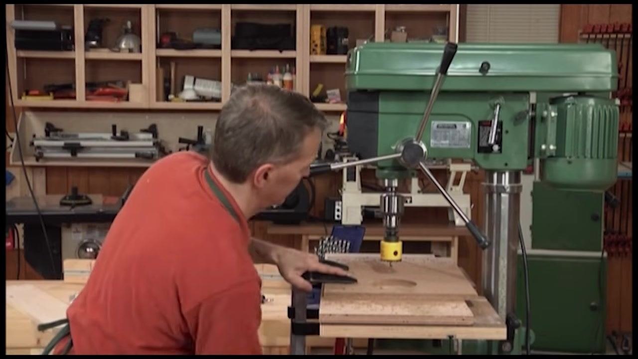 Drill press operating with a yellow hole saw, with the workpiece clamped and the machine set to slow speed