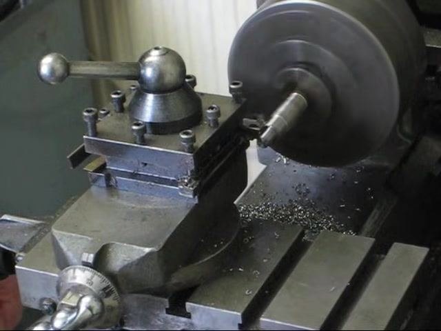 Lathe running with the workpiece spinning in the chuck, cutting tool retracted, and metal chips visible on the bed.