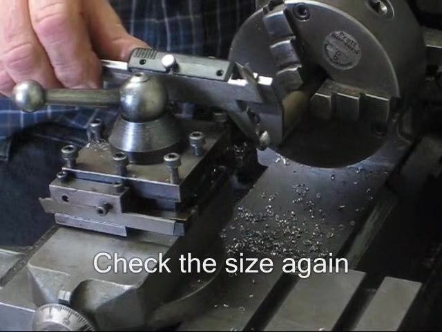 Operator measuring the workpiece diameter with a digital caliper while it remains mounted in the lathe, with metal chips visible on the bed.