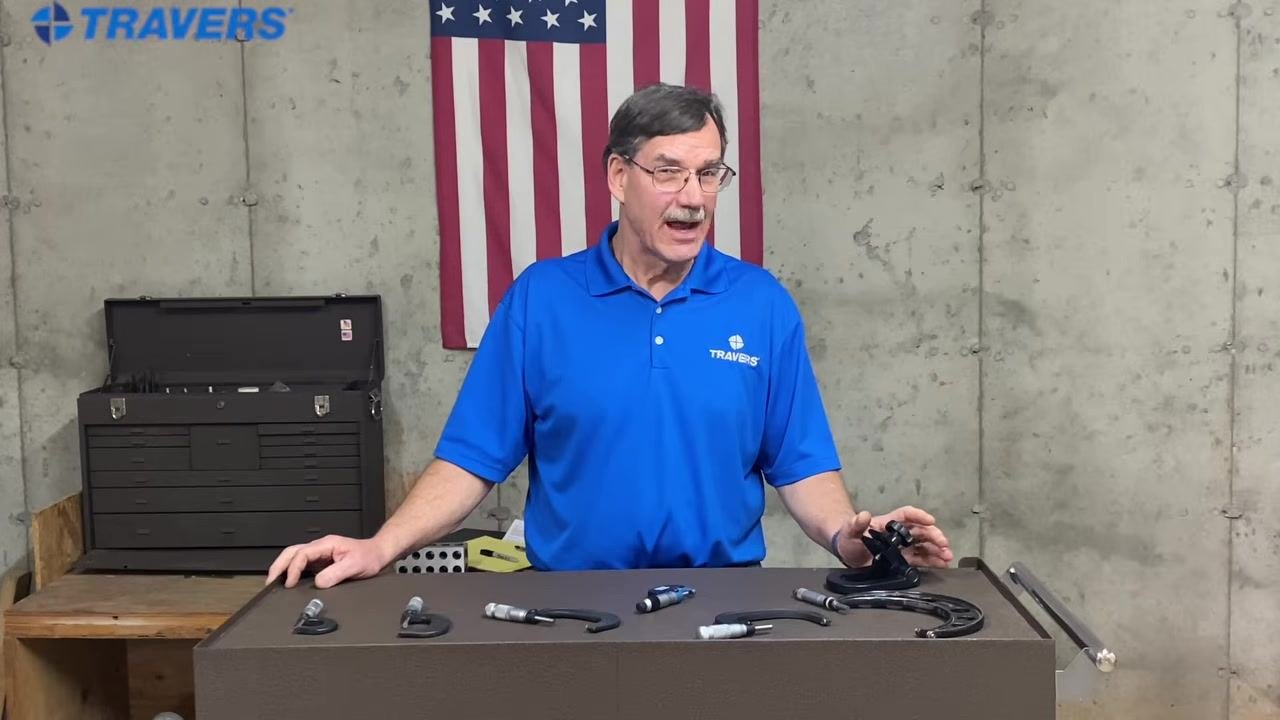 Instructor in blue TRAVERS shirt behind workbench with micrometers, American flag and toolbox in background