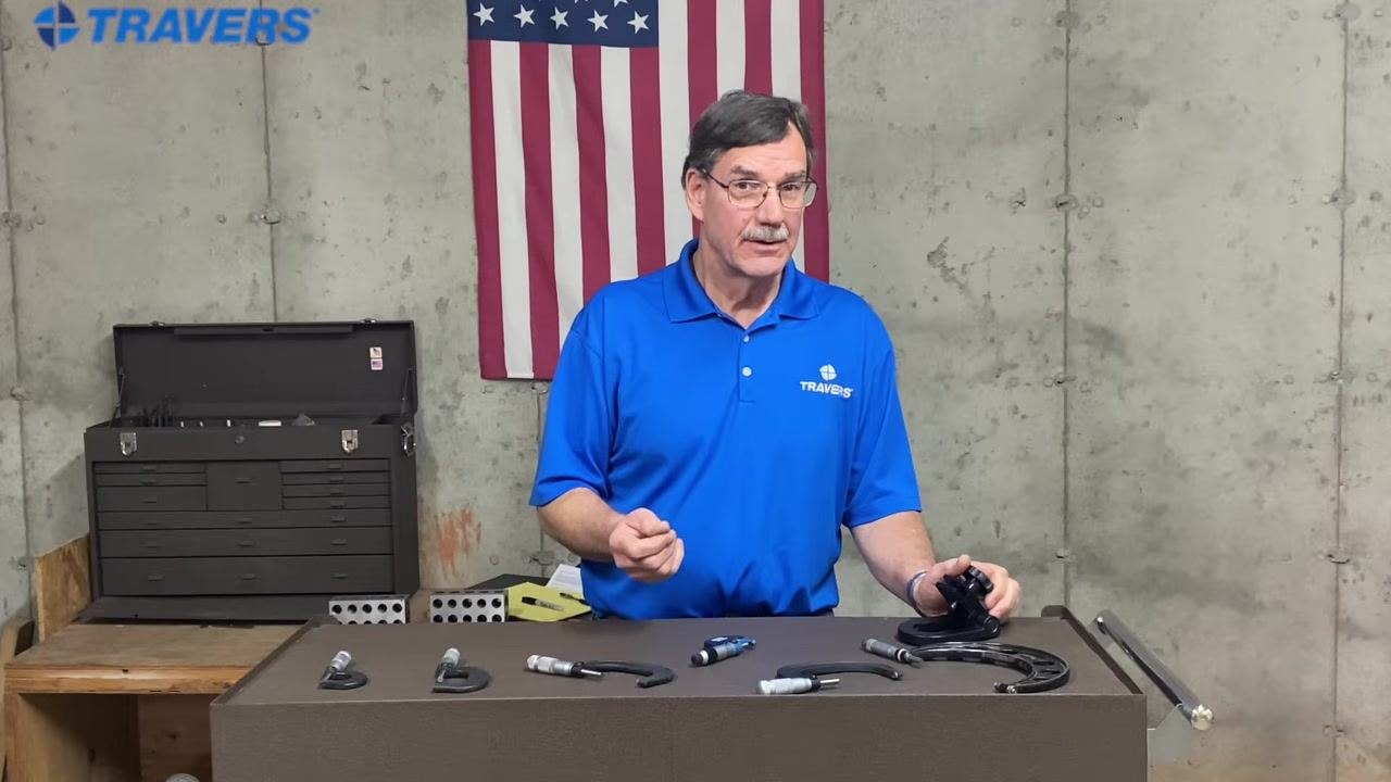 Instructor gesturing with left hand, holding micrometer in right, workbench with micrometers, American flag and toolbox in background