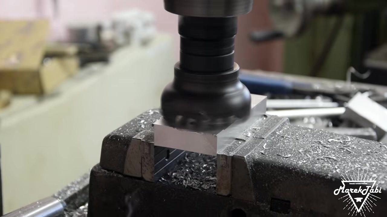 An end mill positioned above the billet, actively machining a new feature near a previously drilled hole, with metal shavings visible on the billet and vise.