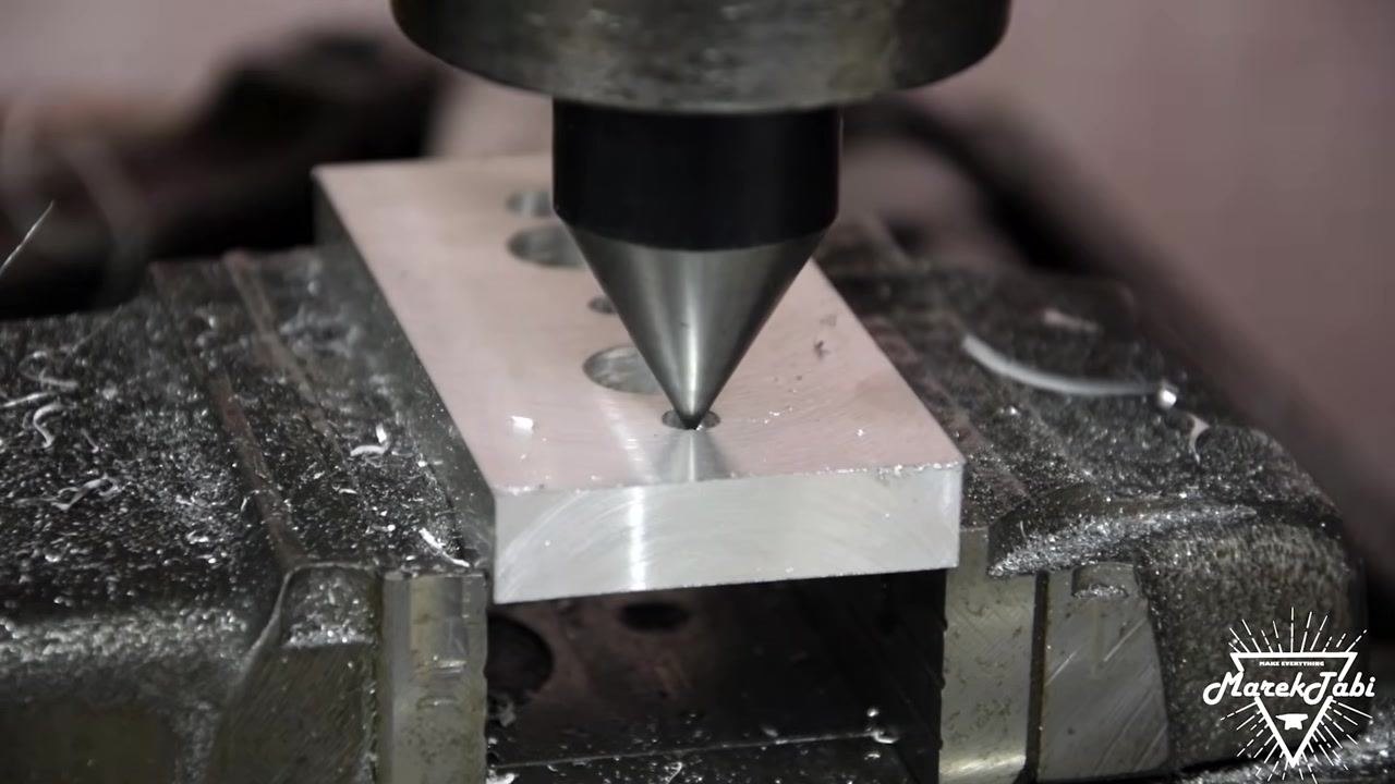 A chamfering tool engaged in a drilled hole on the billet, removing sharp edges and burrs, with multiple holes visible on the billet surface.