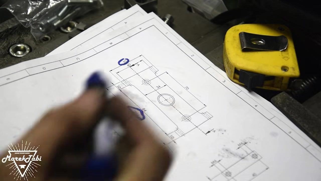 A metal workpiece clamped in a vise with a center drill tool positioned above a marked hole. The work area is covered in metal shavings, and the part is labeled "12" in blue marker.