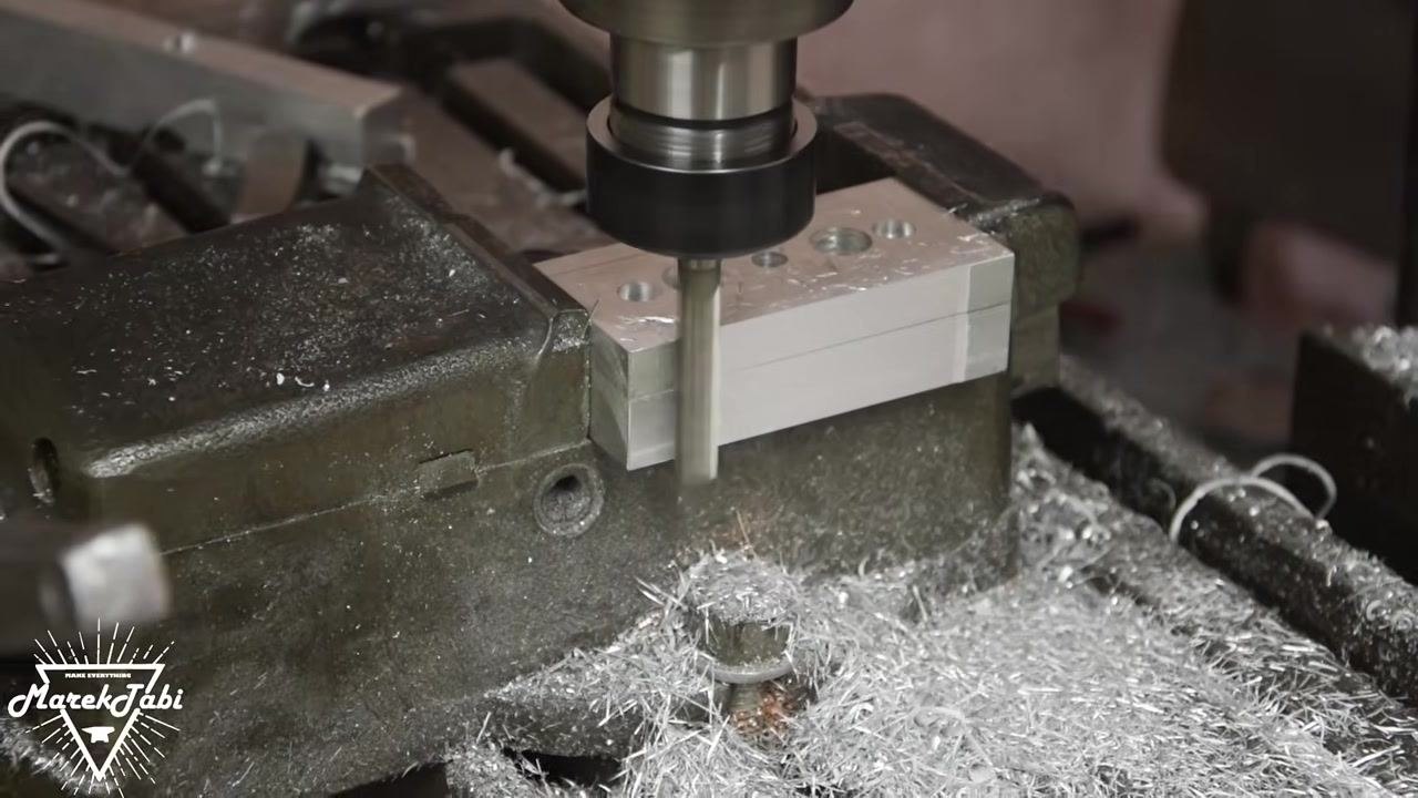A twist drill bit actively drilling into the center hole of a metal workpiece clamped in a vise, with metal shavings visible around the work area.