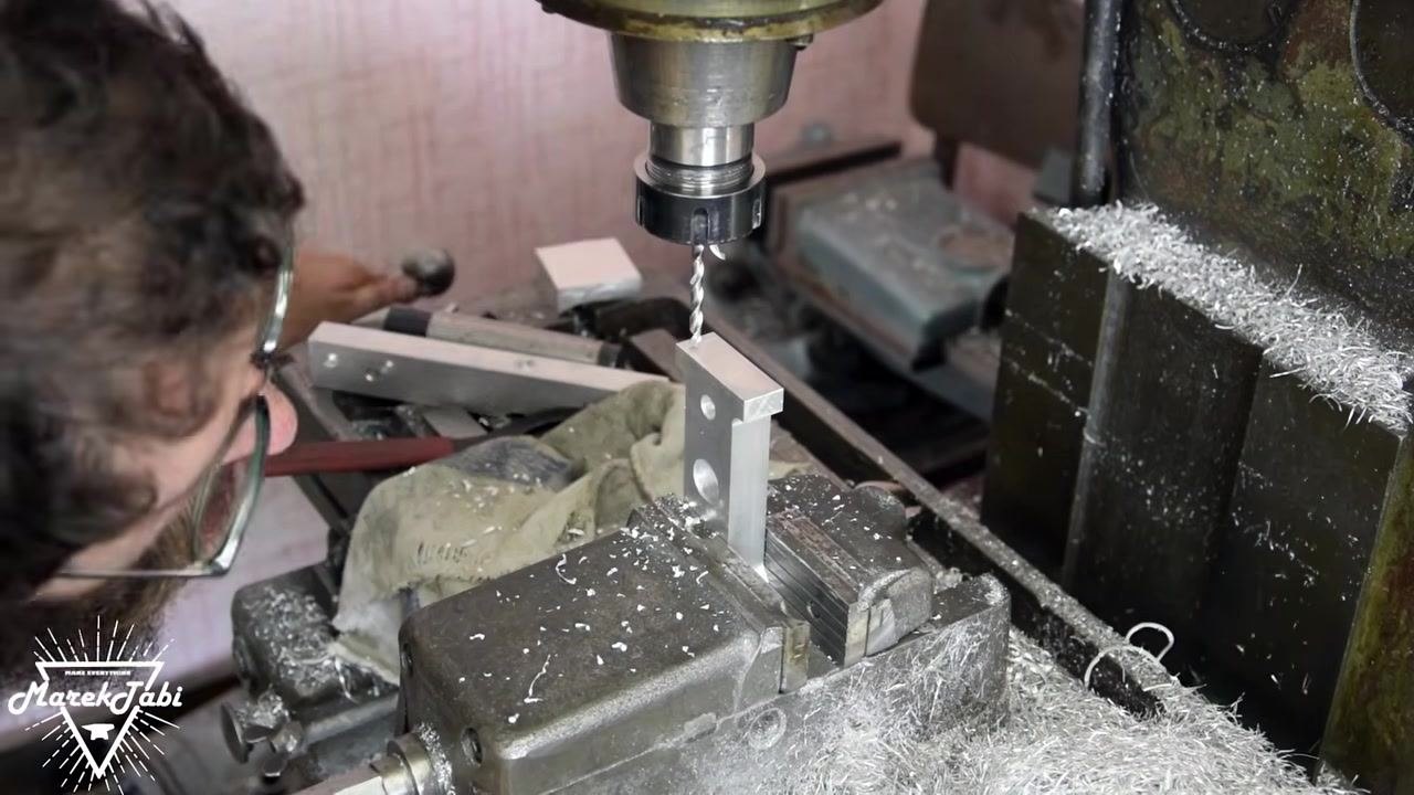A person aligning a drill bit above a vertically clamped metal workpiece in a milling machine vise. The work area is covered in metal shavings, and the operator is adjusting the machine.