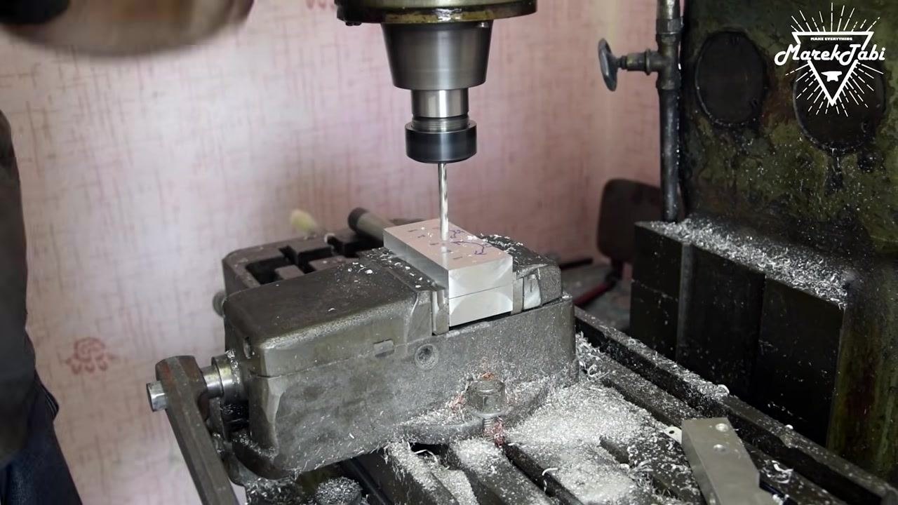 An operator closely aligning a center drill with a marked position on a billet clamped in a milling machine vise, with blue ink marks and arrows visible on the billet.