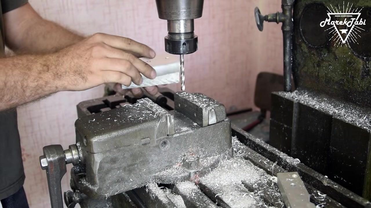 A twist drill bit actively drilling into the billet at a marked location, with metal chips swirling around it and blue ink markings visible on the billet.