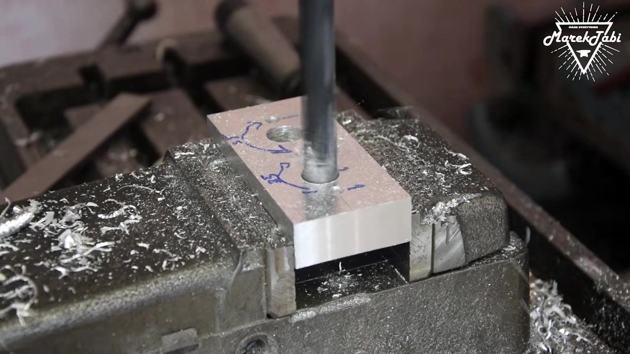 An operator handling the billet after drilling, inspecting the holes, with the vise and machine bed covered in metal shavings.