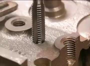 A tap being carefully aligned and started into a drilled hole in a metal workpiece, with several threaded and unthreaded holes visible on the workpiece
