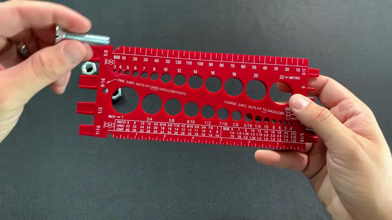 A hand holds a slotted flathead machine screw above a red combination thread gauge displaying multiple diameter holes labeled in metric (MM) and inch sizes, with a nut visible on the table.